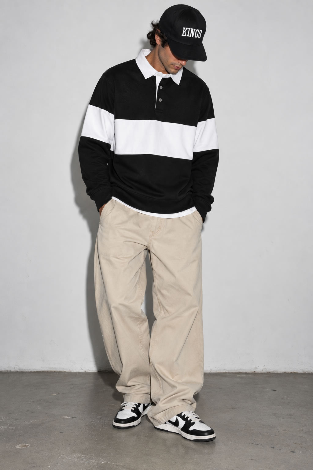 A person wearing a Black And White Striped Men’s Rugby Pullover Shirt, beige pants, black and white sneakers, and a black baseball cap with 'KINGS' on the front, standing against a plain light gray wall.