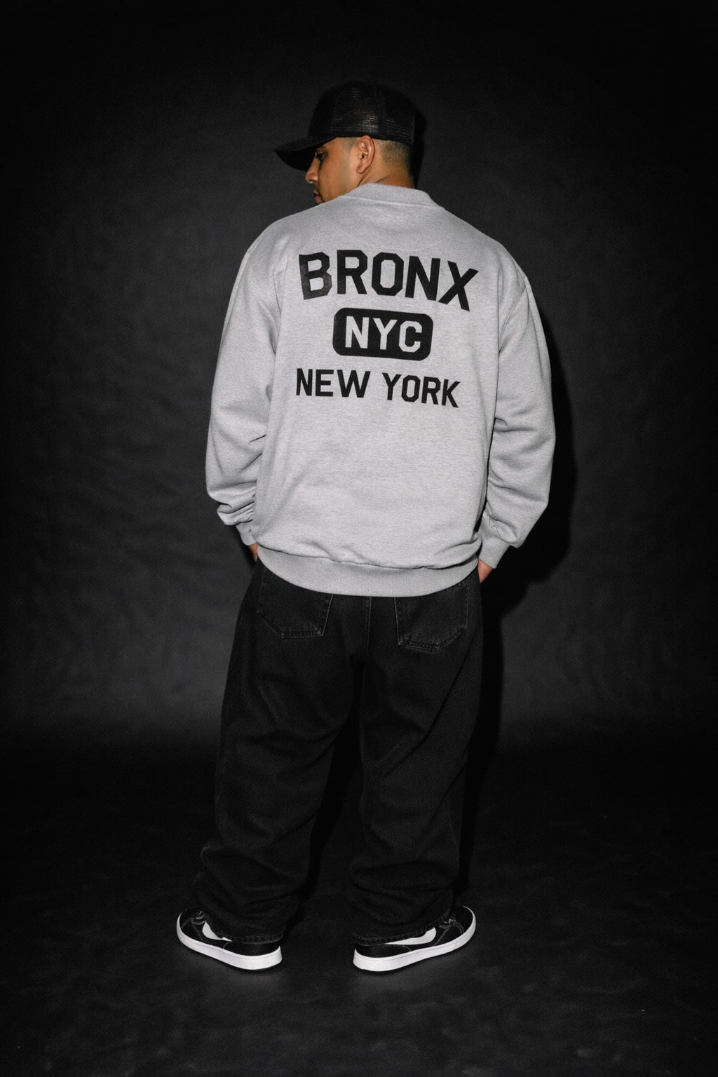 Person wearing a gray bomber jacket with 'Bronx NYC New York' text on the back against a dark background