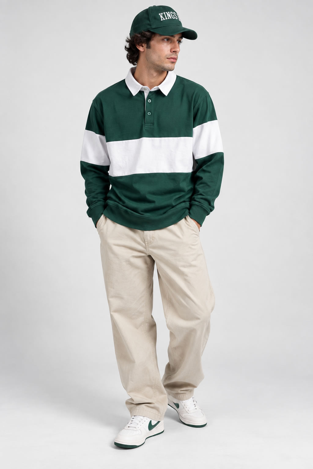 A man modeling a Green And White Striped Men’s Rugby Pullover Shirt, paired with beige pants, white sneakers, and a green baseball cap, against a light gray studio background.