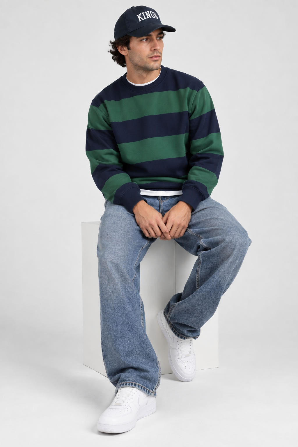 A man wearing a navy and green horizontally striped crewneck sweatshirt, blue jeans, white sneakers, and a baseball cap with 'KINGS' on the front, sitting on a white block against a light gray background.