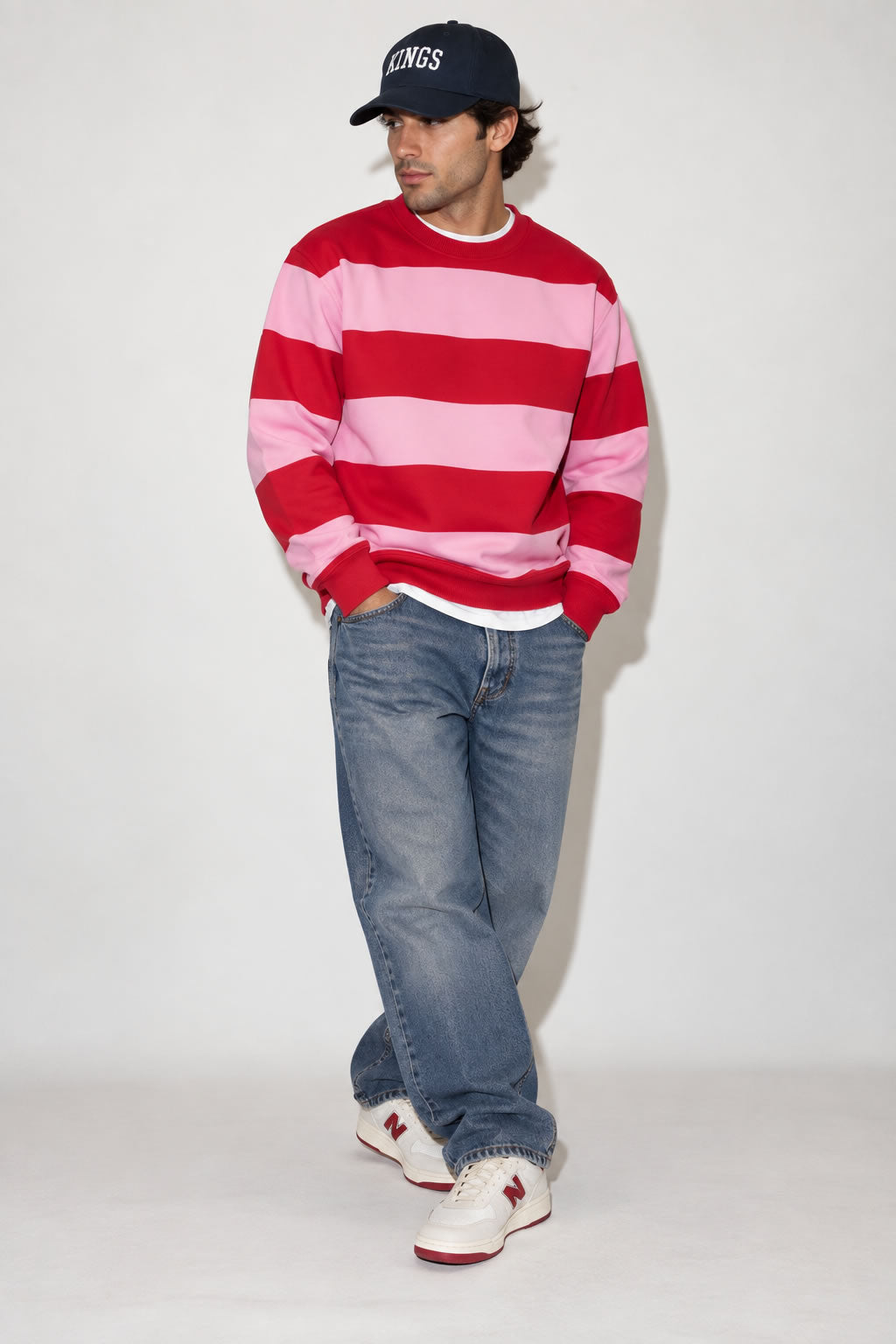 A man wearing a red and pink striped long-sleeve sweatshirt, blue jeans, white New Balance sneakers, and a black baseball cap with 'KINGS' written on it, standing against a plain light gray studio background.