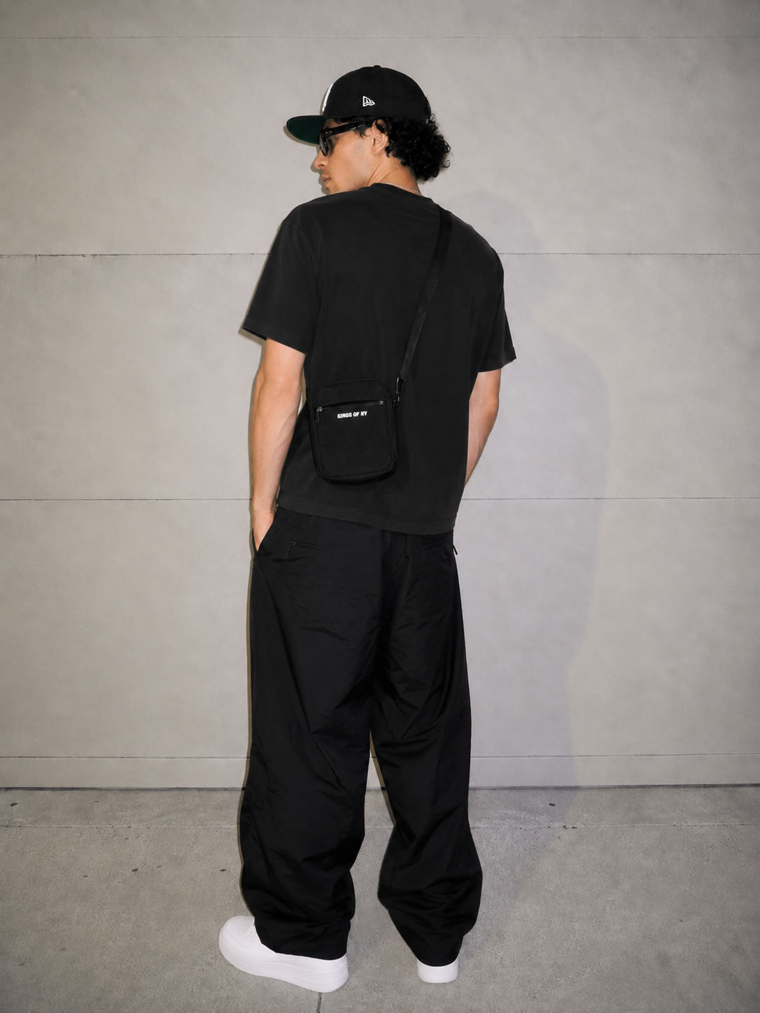 A person shown from the back wearing a black t-shirt, black baggy pants, a black baseball cap, and white sneakers, while wearing a small black crossbody bag with a white logo. The background is a minimalist gray concrete wall.