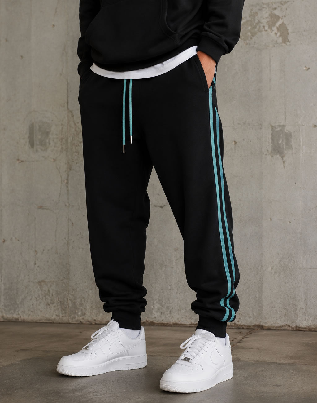 A person wearing black jogger sweatpants with light blue side stripes and matching drawstrings, paired with a black hoodie and white sneakers, standing against a textured concrete wall.