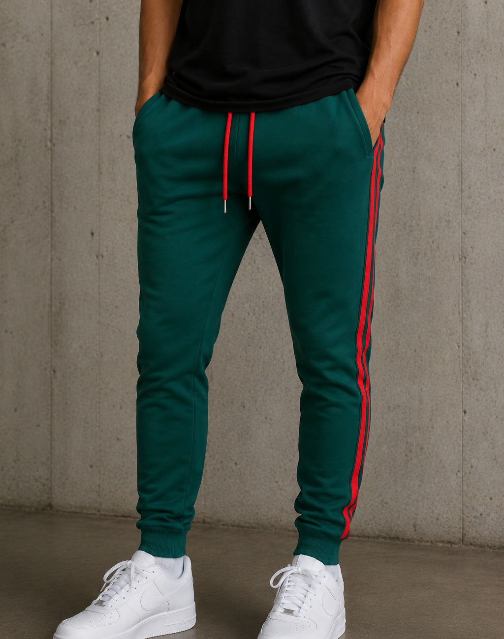 A person wearing dark green jogger sweatpants with red side stripes and a red drawstring, paired with a black top and white sneakers, standing against a concrete wall.