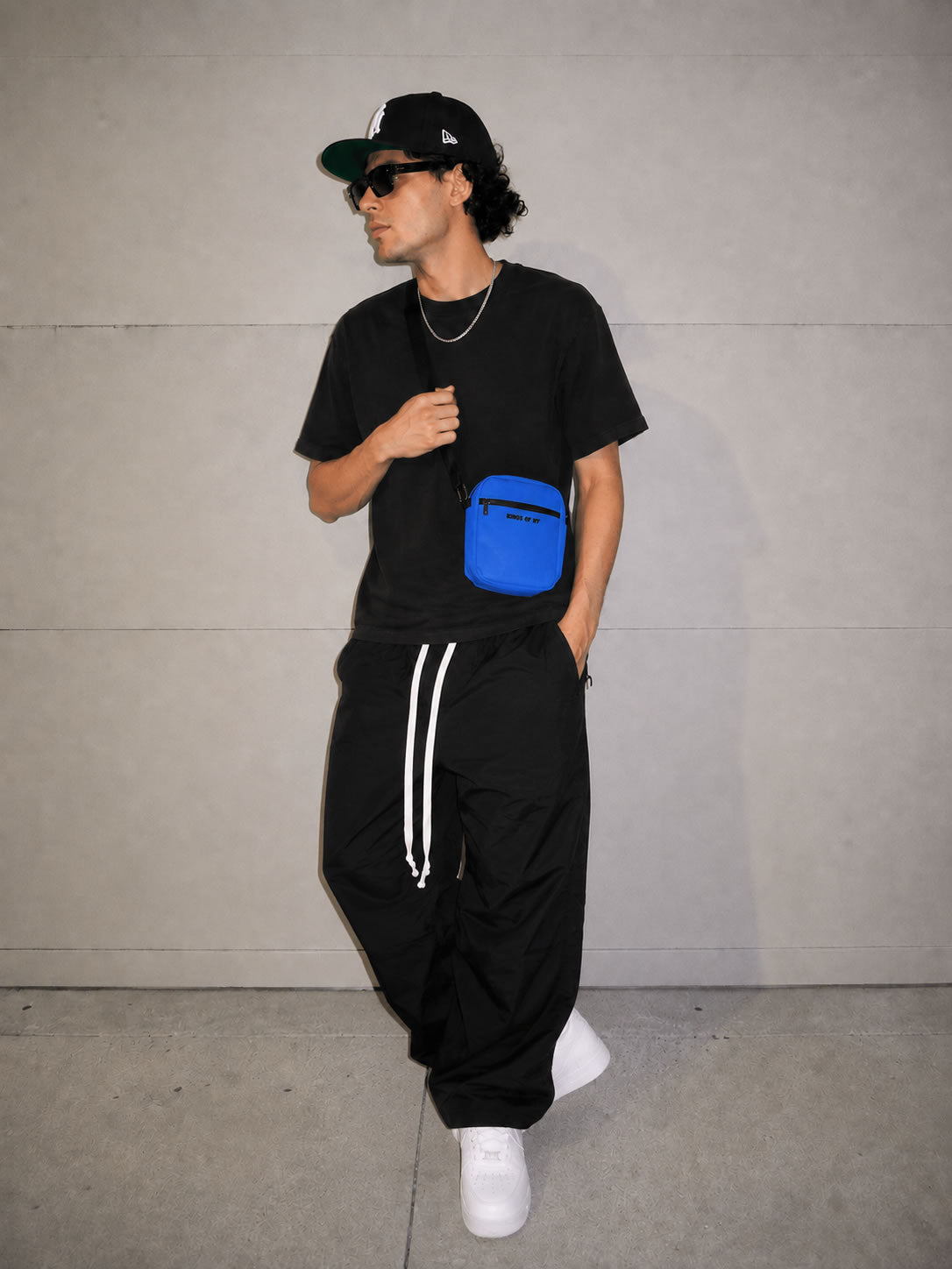 A model wearing a black t-shirt, black sweatpants with a kings of ny royal blue crossbody bag against a concrete wall.
