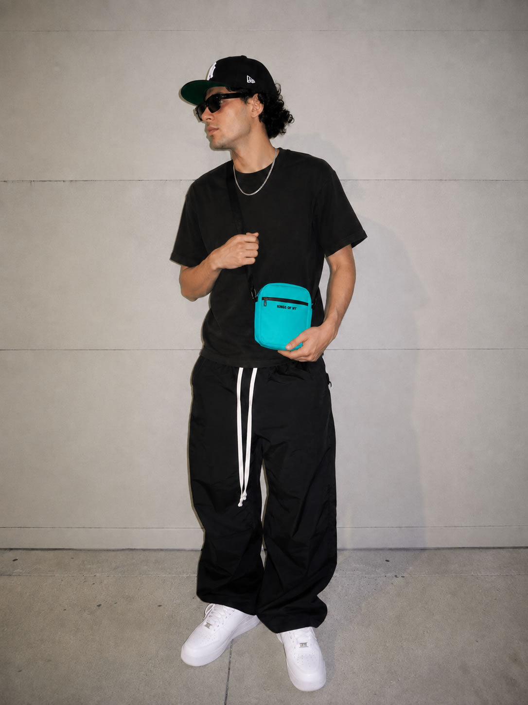 A man wearing a black t-shirt, black sweatpants with white side stripes, a black baseball cap, and white sneakers, holding a bright turquoise crossbody bag against a light gray concrete wall.